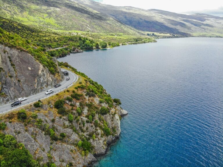 Aerial photograph capturing a scenic road by a lake in New Zealand with lush greenery and mountains in the background.